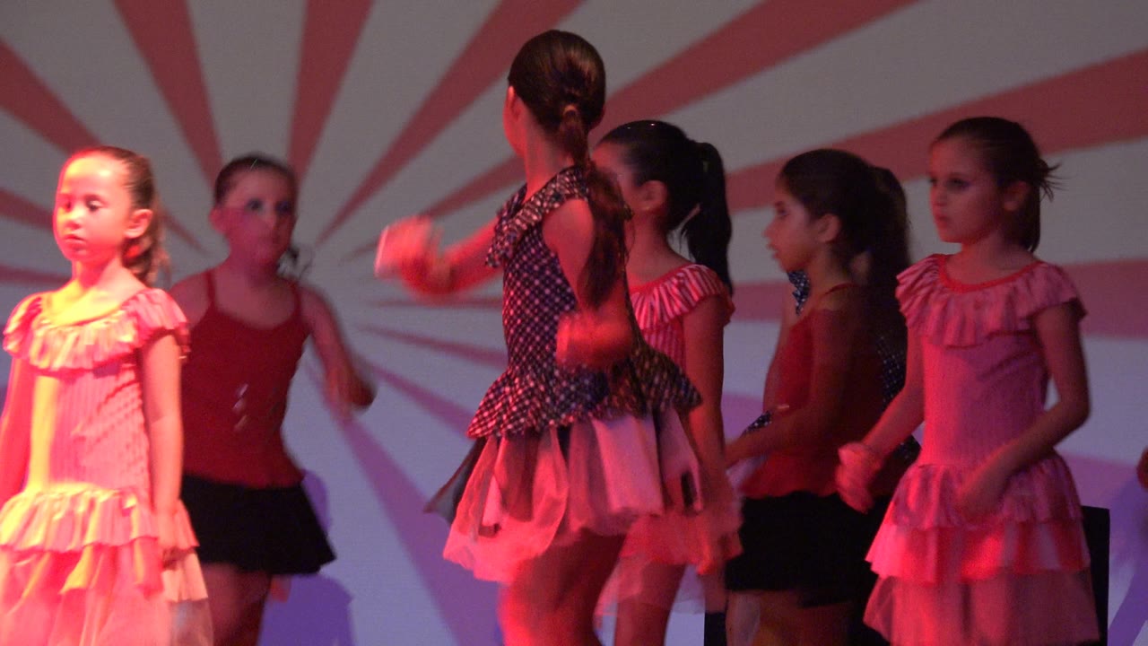 Young dancers in San Jose del Cabo BCS Mexico shift positions on a red-lit stage, with a girl in a polka-dot top and tulle skirt centered near the front.
