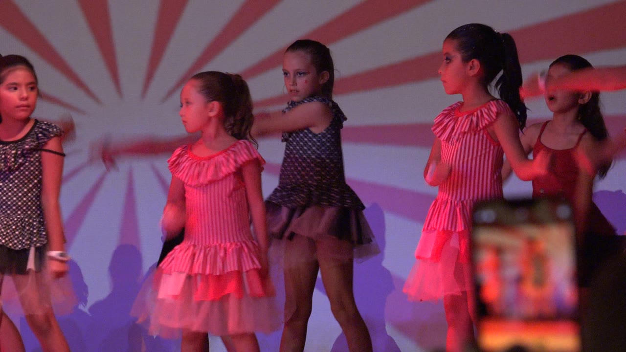Young girls perform on a stage in San Jose del Cabo, Mexico, wearing pink and dark patterned dresses against a red and white sunburst backdrop.