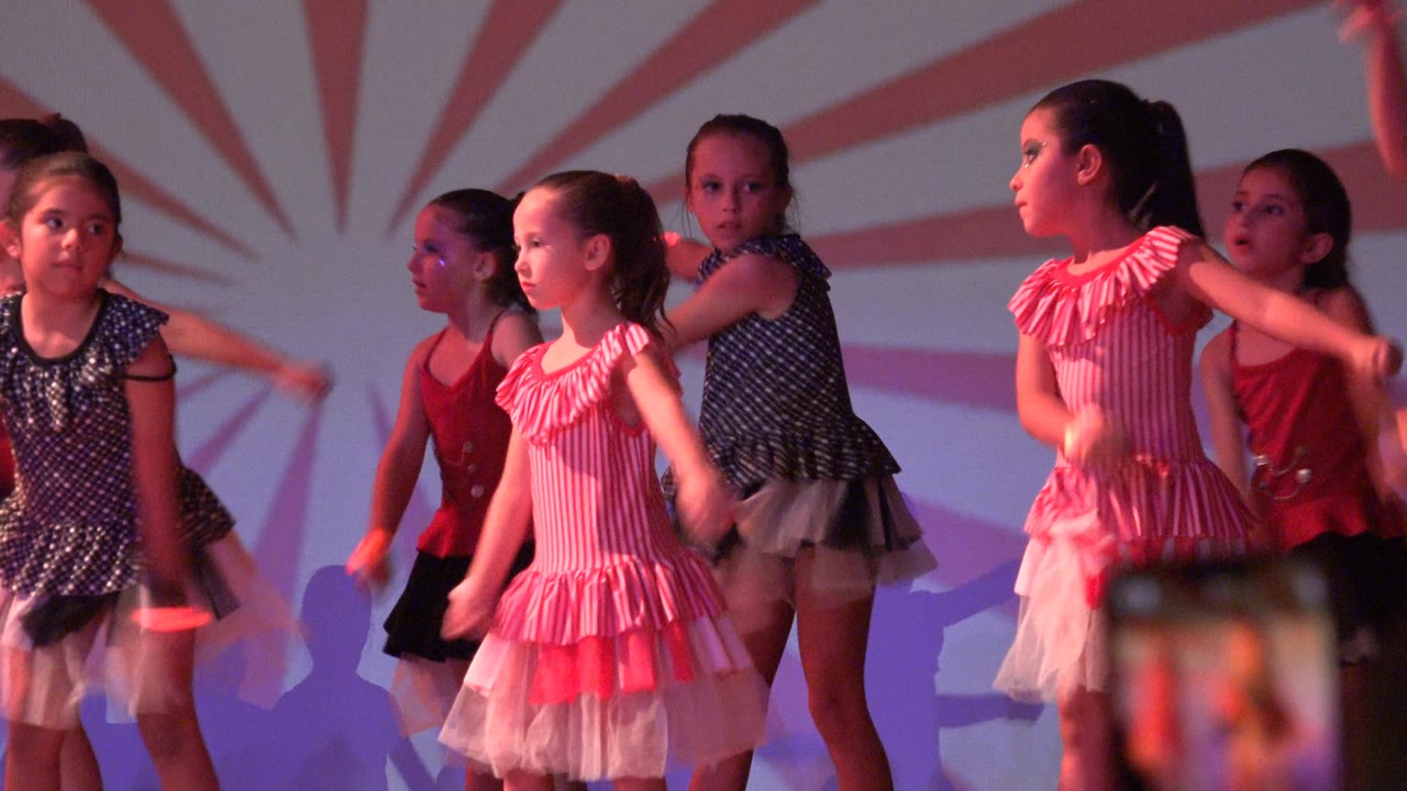 Girls in dresses continue a stage dance while a phone appears in the foreground.