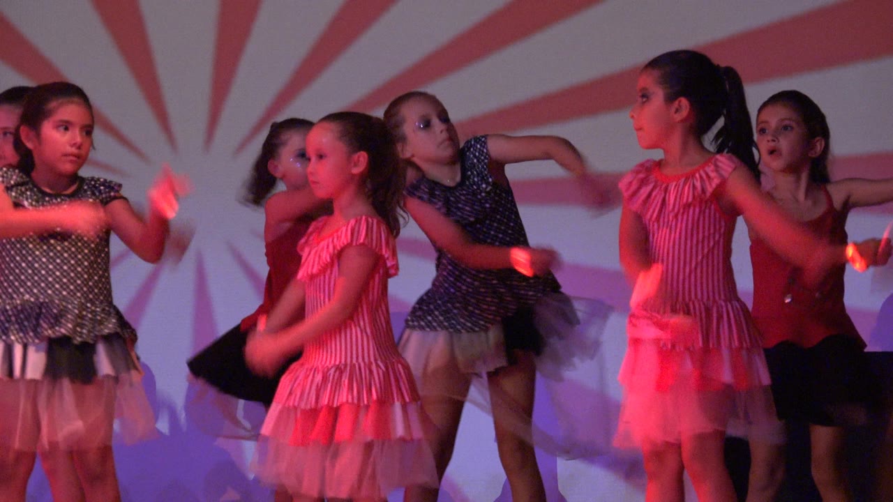 Young girls in pink, red, and dark dresses dance in formation in San Jose del Cabo, Mexico, with arms moving under red stage light.