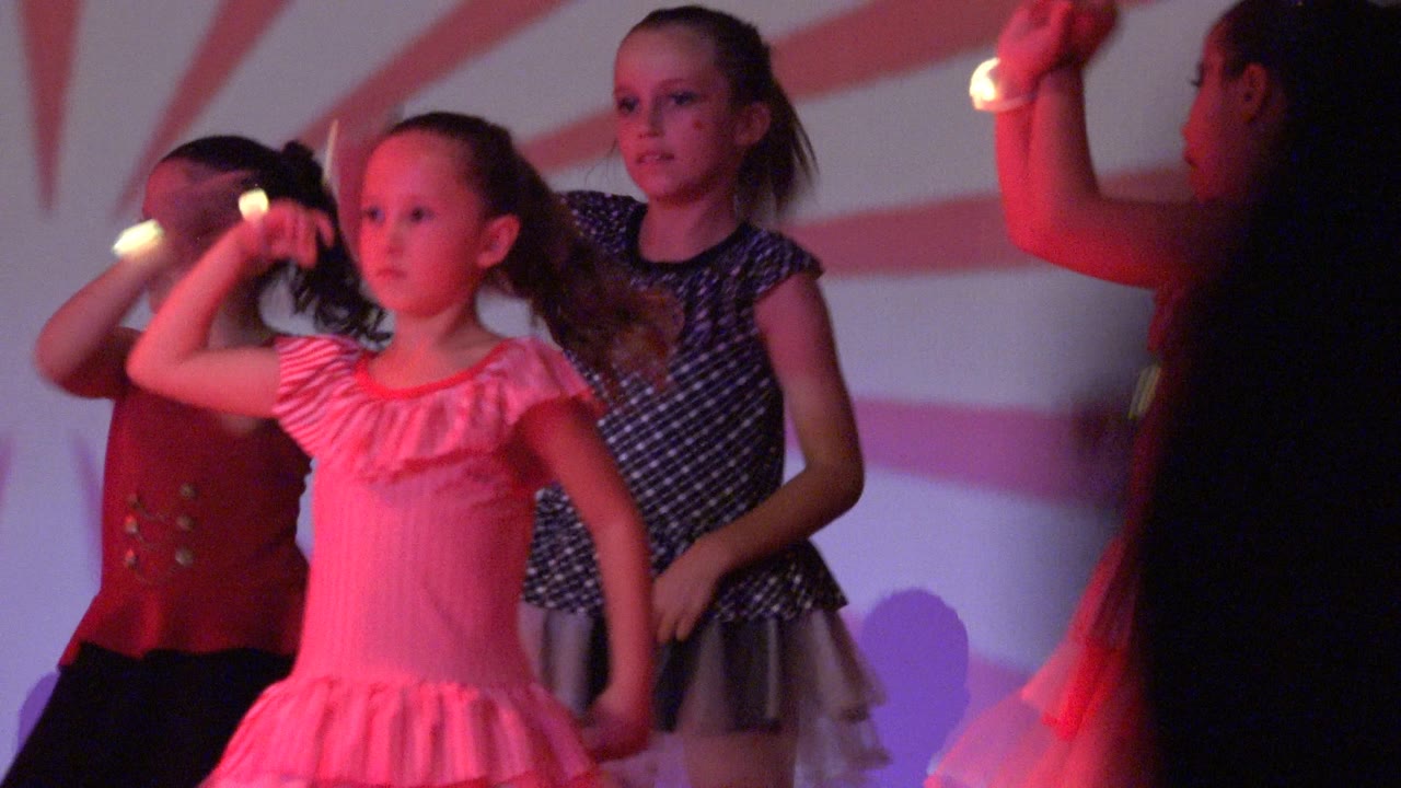 Young girls perform on a stage in San Jose Del Cabo BCS Mexico, with one child raising a glowing stick and red stripes projected across the backdrop.