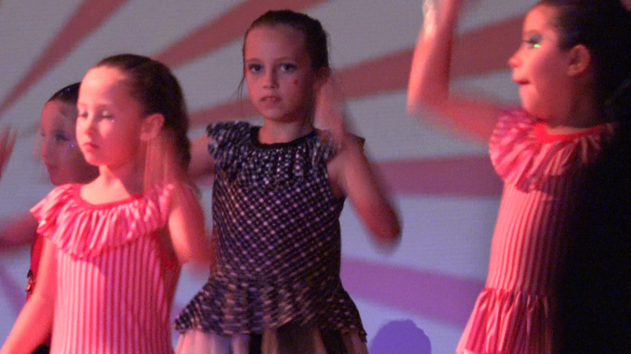 Children in dresses move their arms during a dance routine.