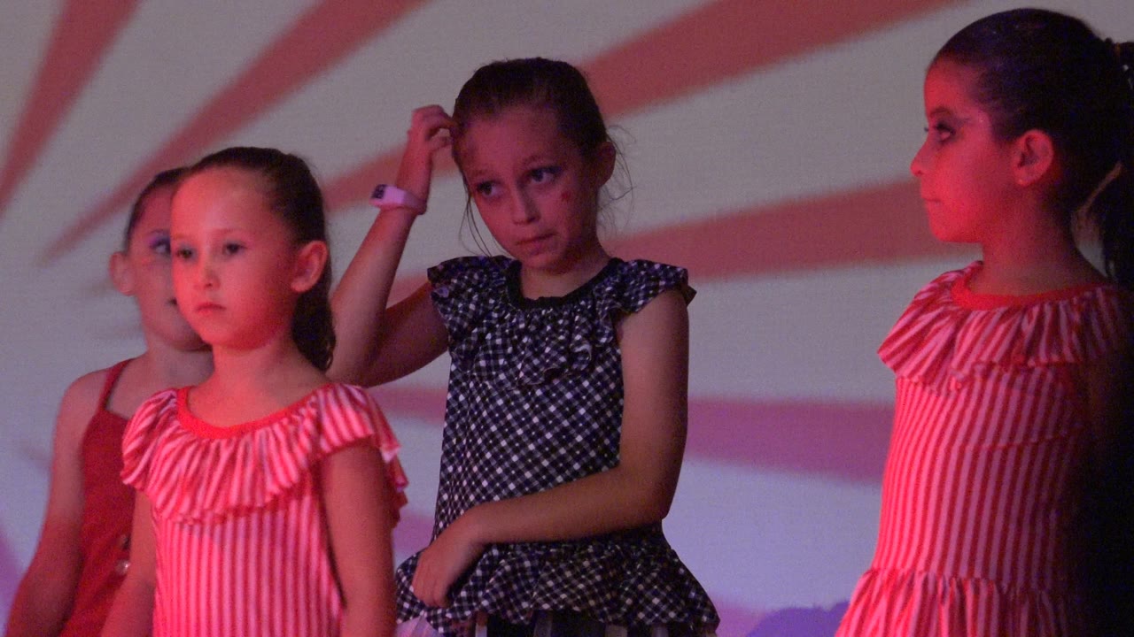Young girls perform on a stage in San Jose Del Cabo BCS Mexico, with pink-red lighting, a sunburst backdrop, and one child touching her hair.