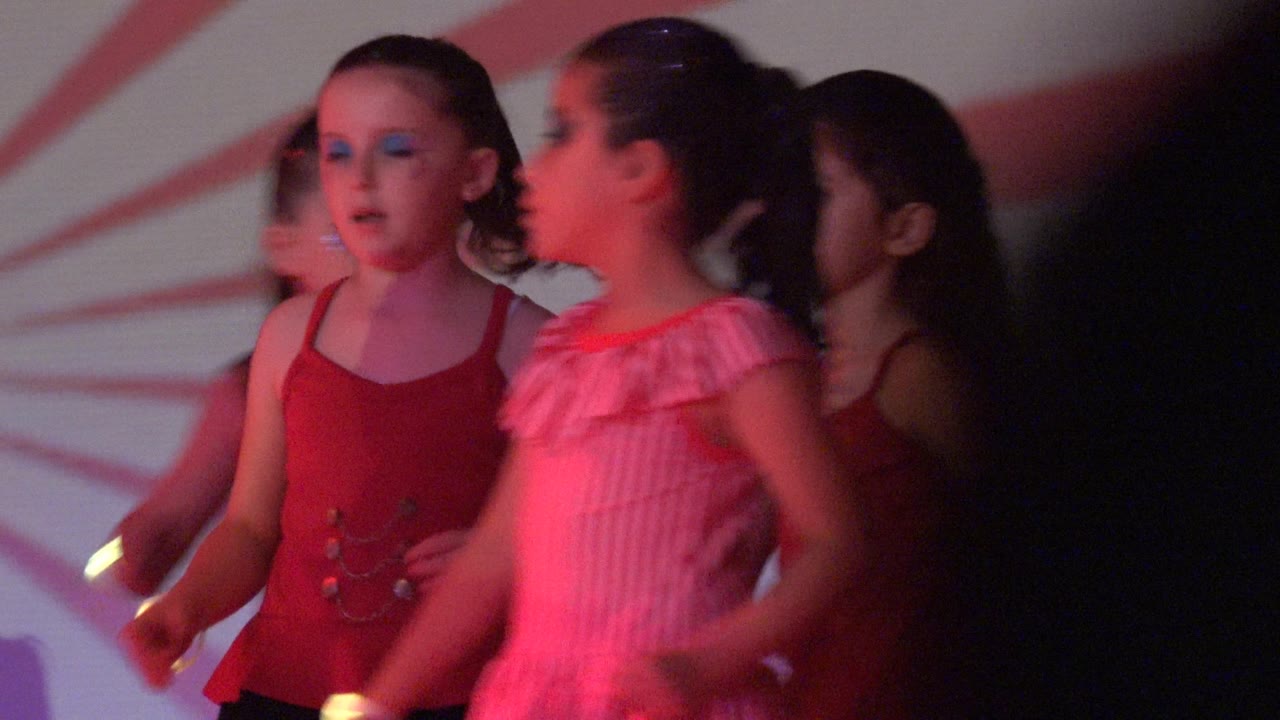 Girls in red and pink outfits move across the stage in San Jose Del Cabo BCS Mexico, with small glow sticks visible against the red-pink wash.