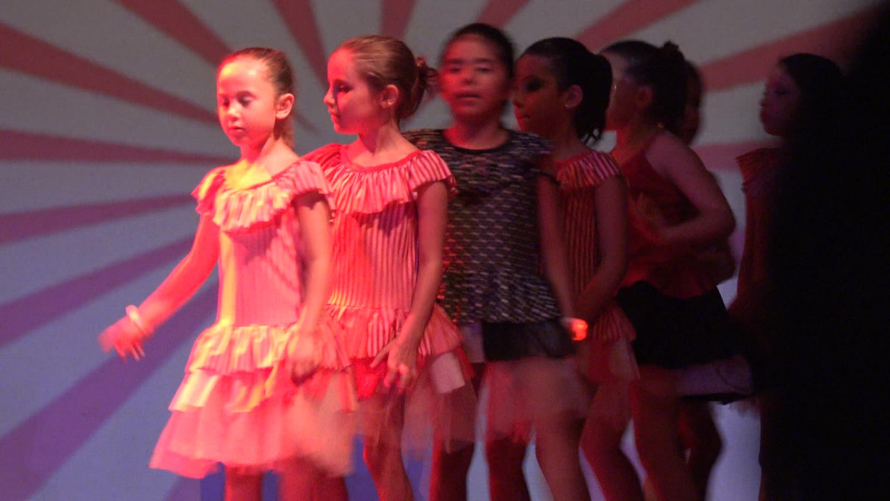 Young girls in San Jose del Cabo, Mexico stand on stage in pink, red, and dark dance costumes under red lighting with a sunburst backdrop.