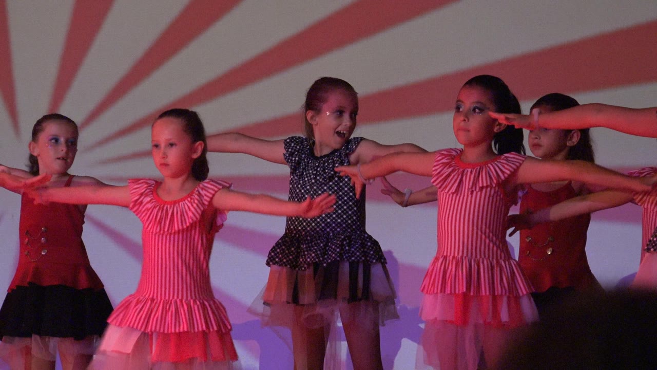 Young girls in pink, red, and black dance costumes hold extended arm positions on a stage in San Jose Del Cabo BCS Mexico, with a red and white radial backdrop behind them.