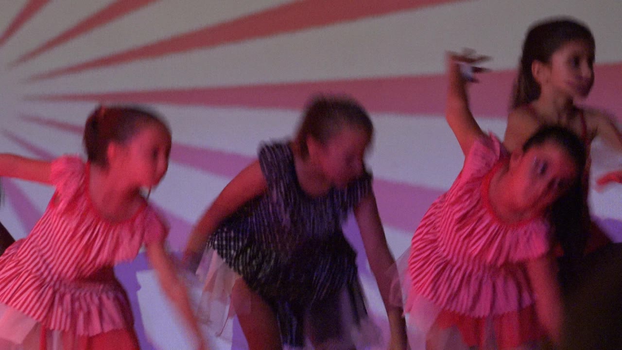 Young girls in pink and dark costumes bend and lift their arms during the performance in San Jose Del Cabo BCS Mexico, with the backdrop pattern radiating behind them.