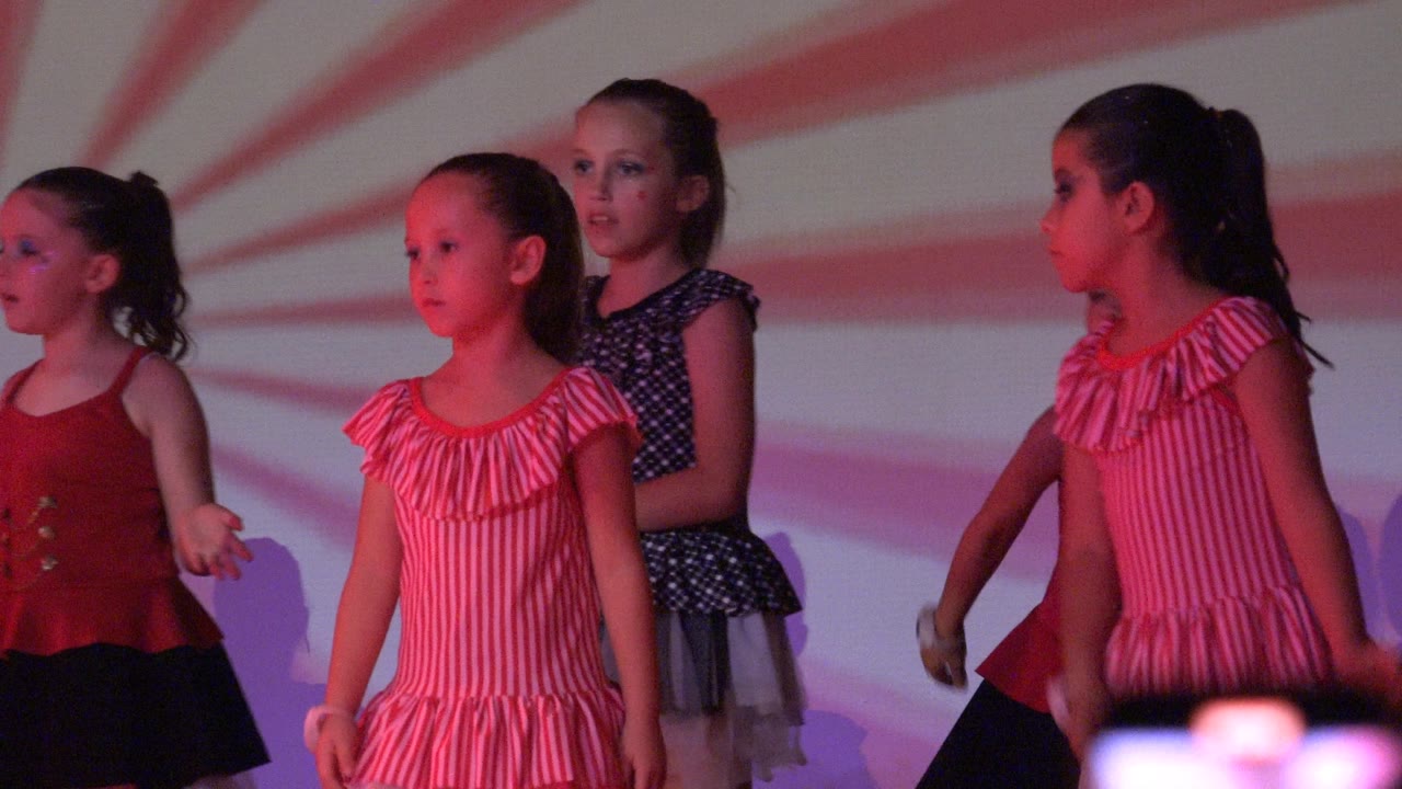 Young girls in red, pink, and dark costumes stand on a stage in San Jose del Cabo, Mexico, with a red striped projection behind them.