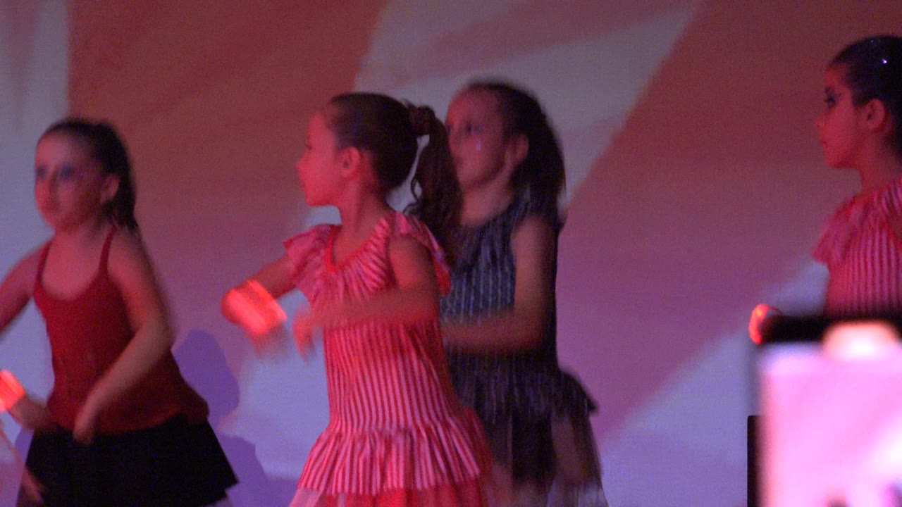 Young girls dance on a San Jose del Cabo stage while holding small glowing objects, with blurred audience phone screens visible along the bottom edge.