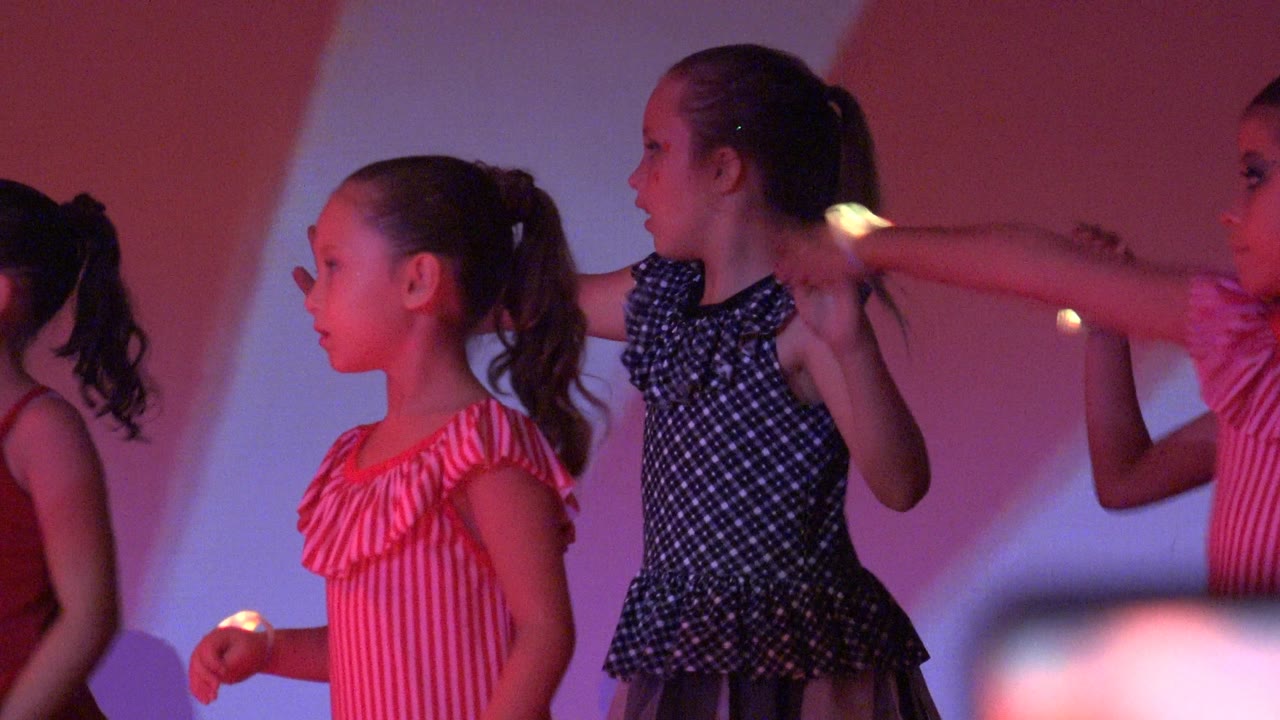 Young girls perform a stage dance in San Jose Del Cabo BCS Mexico, with pink striped dresses, a dark polka dot dress, extended arms, and colored light on the backdrop.
