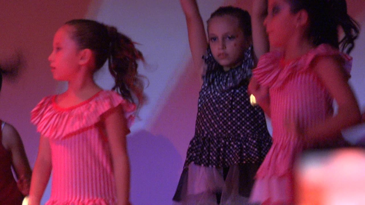 Young girls in San Jose Del Cabo BCS Mexico perform on stage under red and purple lighting, with a pink striped dress prominent at left and a black polka-dot costume near center.