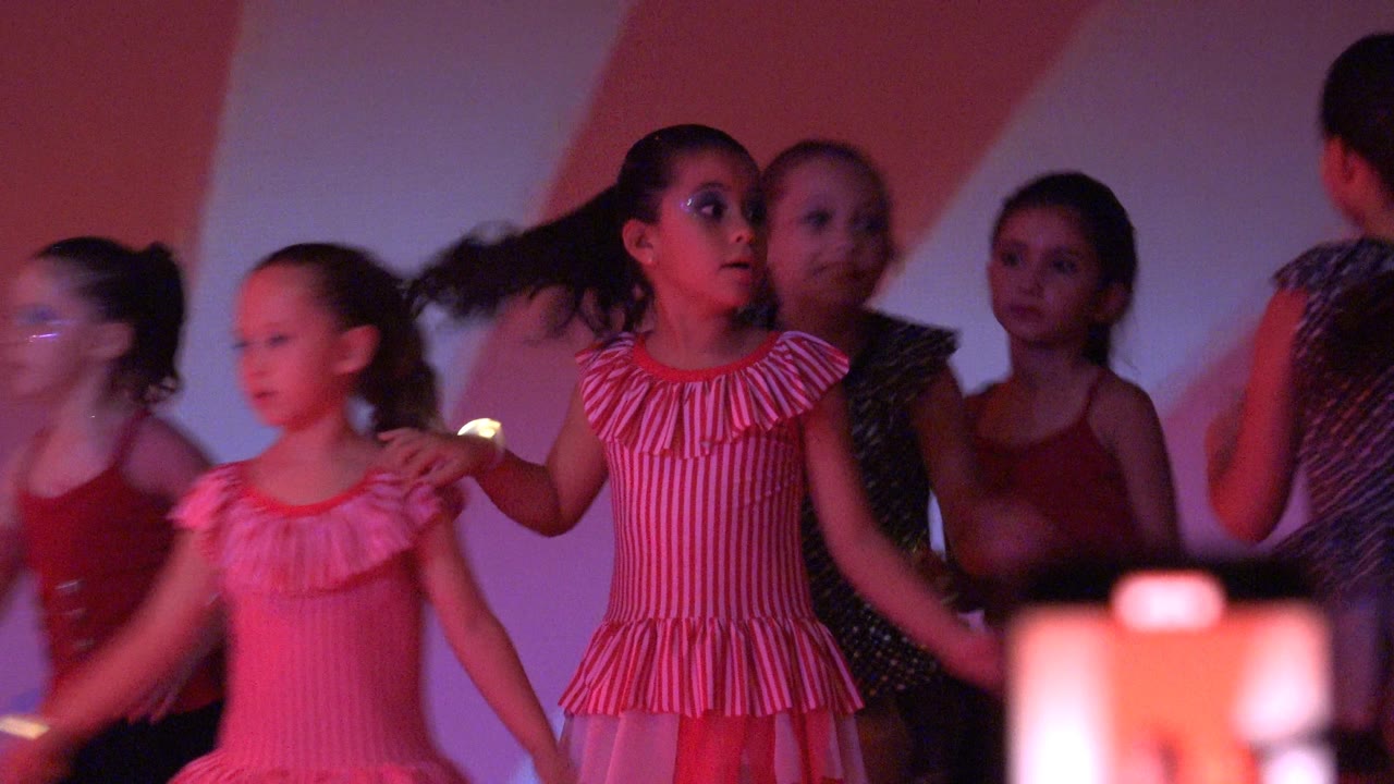 Young girls perform on a stage in San Jose Del Cabo BCS Mexico, with one striped pink dress prominent and a blurred phone visible at the bottom edge.