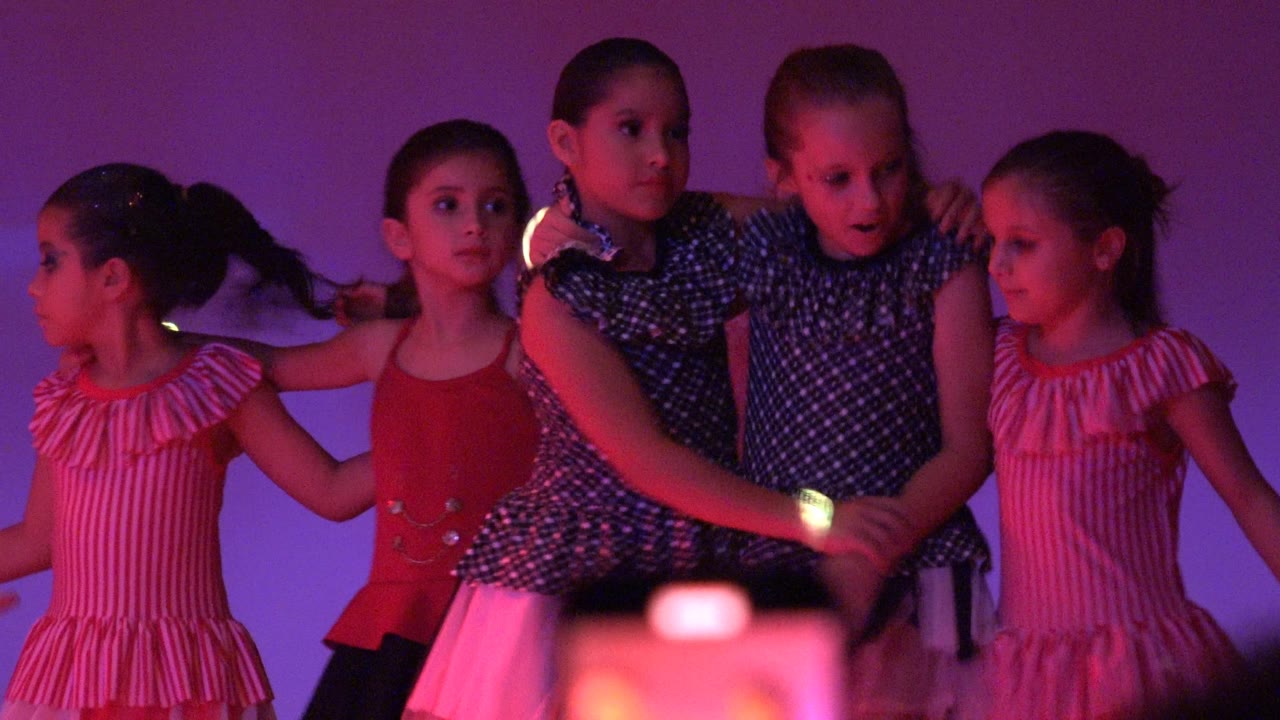 Several young girls in red, pink, and dark patterned dresses stand close together and hold hands during the dance in San Jose Del Cabo BCS Mexico.