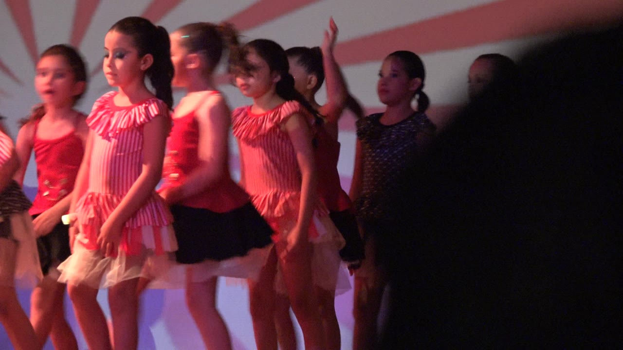 At 0:01 in San Jose Del Cabo BCS Mexico, several girls in red, black, and striped dance outfits stand in a line under red lighting with the sunburst backdrop behind them.