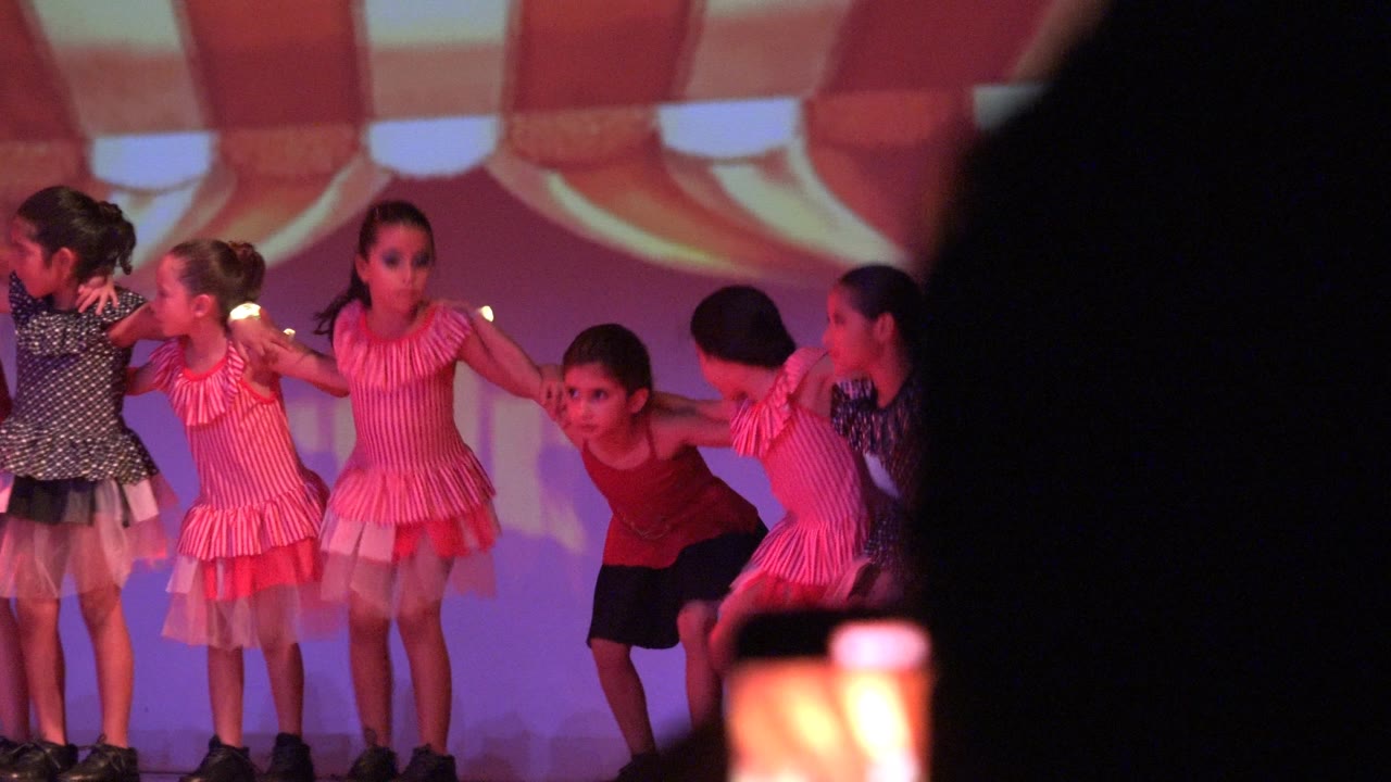 Children perform on a stage in San Jose del Cabo BCS Mexico, with several girls in pink dresses and a child in red bending forward as a dark audience silhouette covers part of the frame.