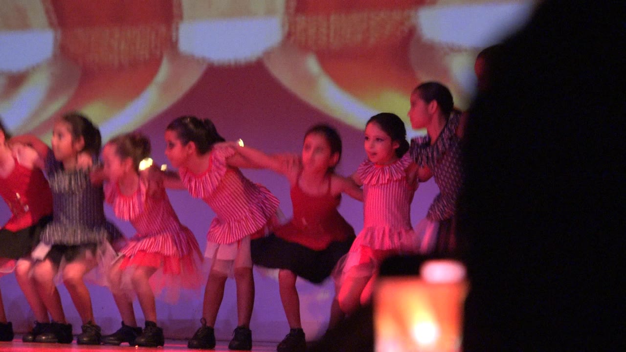 Children in matching pink, red, black, and white outfits crouch and lean in unison on a stage in San Jose del Cabo BCS Mexico while audience foreground shapes partially block the lower frame.