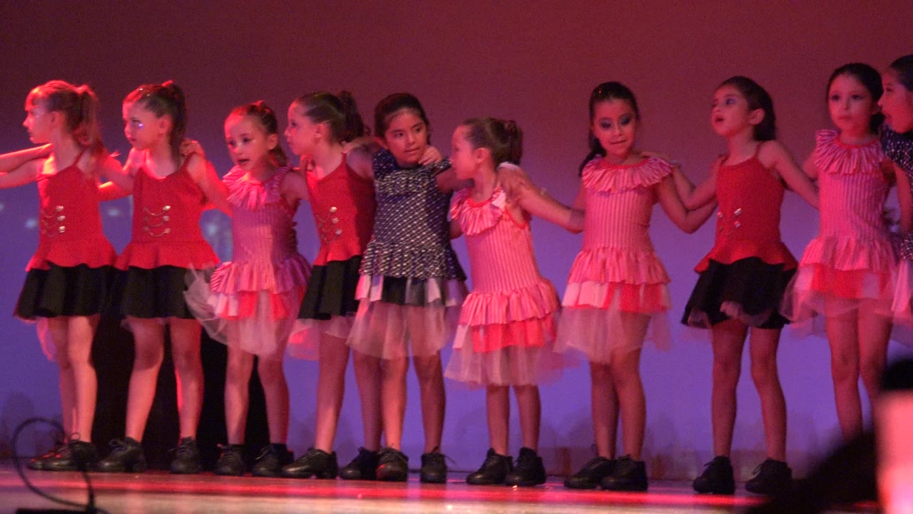 Young girls in red, pink, black, and white dance costumes stand shoulder to shoulder on a stage in San Jose Del Cabo BCS Mexico under purple and red light.