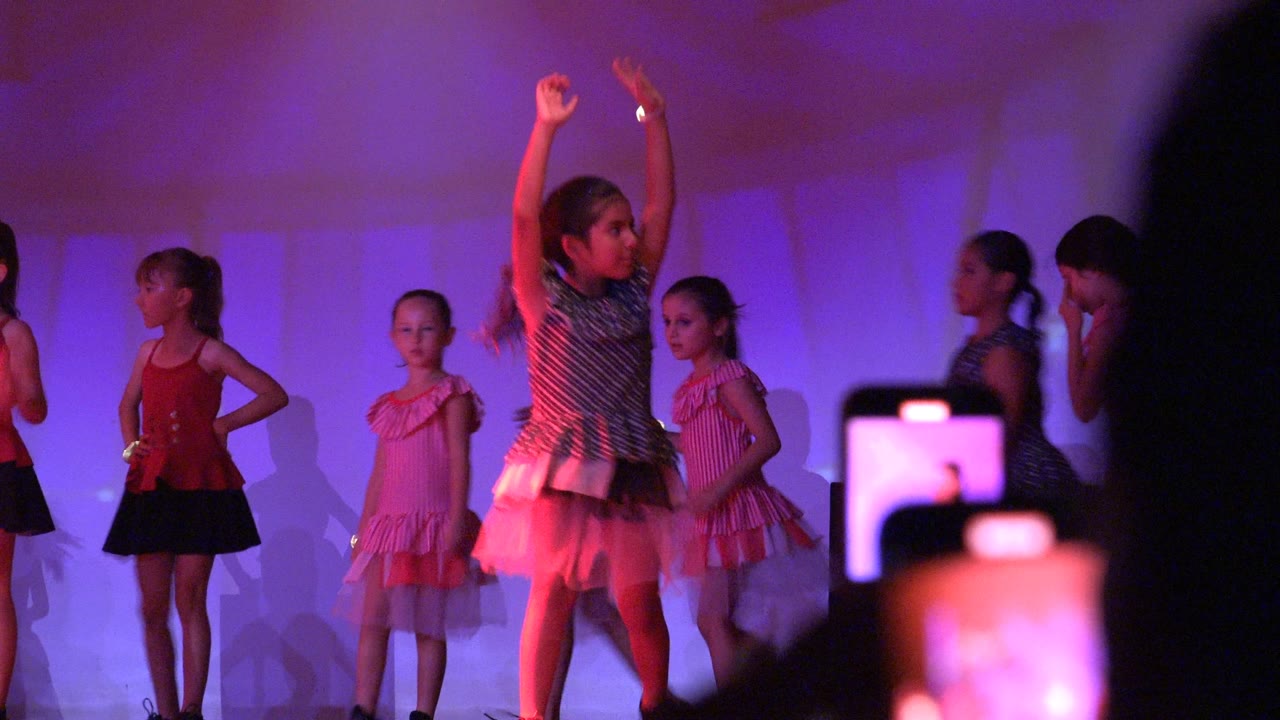Young girls perform a stage dance in San Jose Del Cabo BCS Mexico, with one dancer's arms raised and an audience smartphone visible at the right edge.