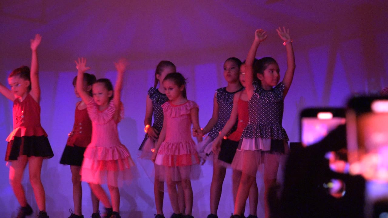 Girls in pink, red, and dark costumes raise their arms during the routine in San Jose Del Cabo BCS Mexico while audience phones appear along the lower right foreground.