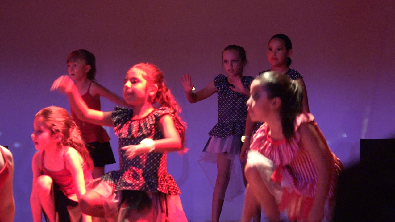 Young girls perform a synchronized stage dance in San Jose del Cabo, Mexico, with one dancer crouched in front and others standing behind under purple and red lighting.
