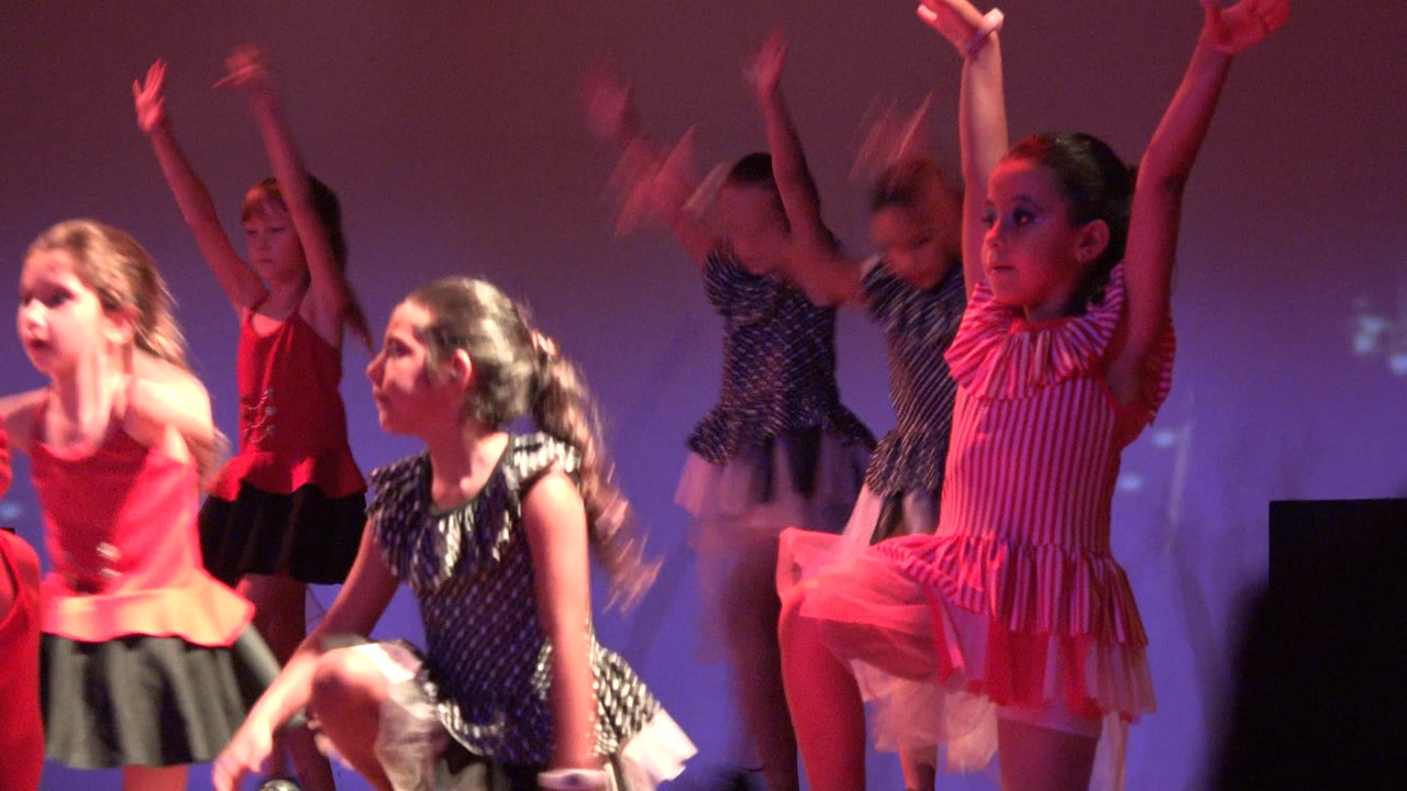 Girls in colorful dance outfits raising arms on stage.