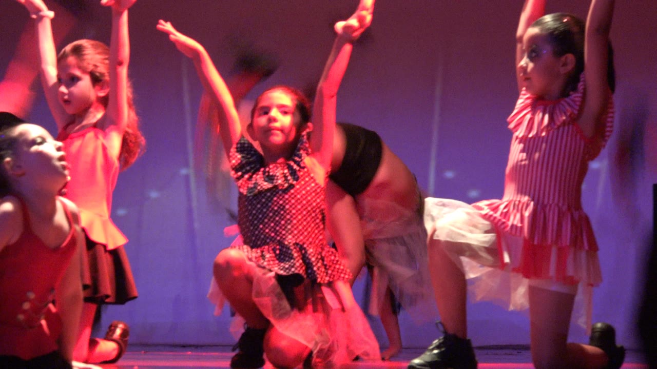 Several young dancers hold high arm positions on a purple-lit stage in San Jose del Cabo, Mexico, while a girl in a polka-dotted dress kneels near the foreground.