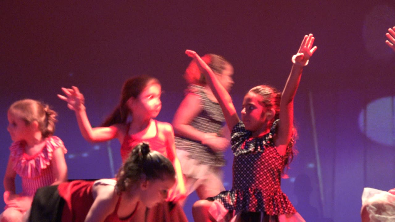 Young girls perform on a stage in San Jose Del Cabo BCS Mexico, with raised arms, ruffled dresses, and pink-purple lighting across the backdrop.