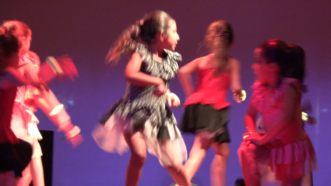 Girls moving across a lit stage in colorful dance outfits.