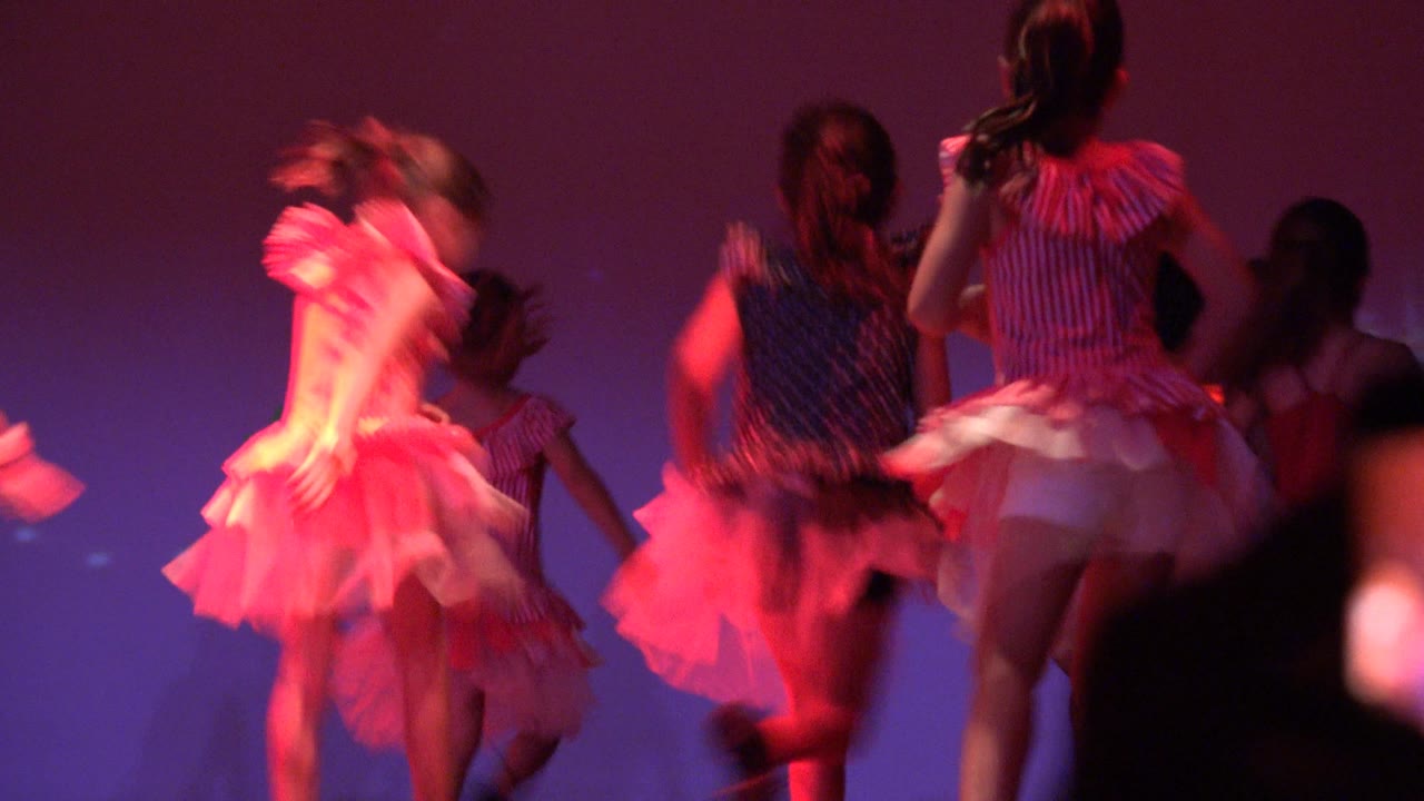 A girl in a dark patterned dress turns near center stage while other children in pink and red outfits move around her in San Jose Del Cabo BCS Mexico.