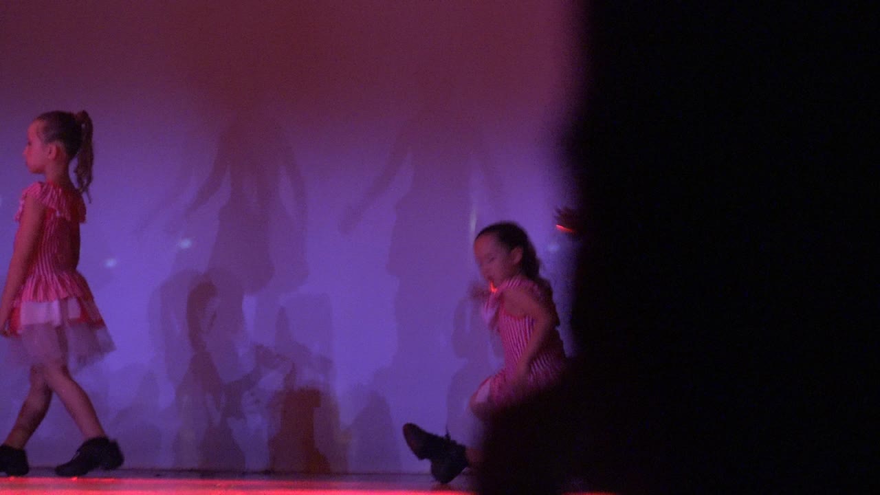 Two young girls in pink dance costumes are visible on a purple-lit stage in San Jose Del Cabo BCS Mexico, with one walking at left, one crouching near center, and a dark foreground shape blocking the right side.
