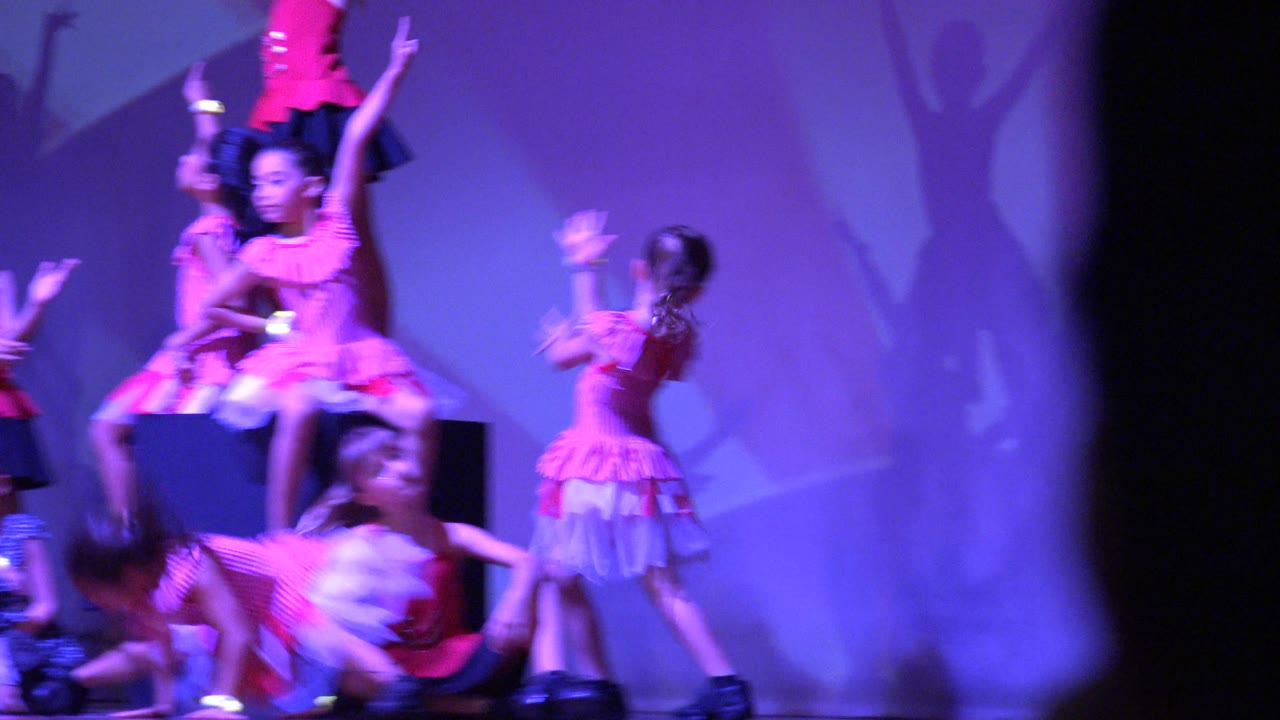 A group of young girls in pink and red outfits pose on a stage in San Jose Del Cabo BCS Mexico, with one dancer on a black platform and large dancer shadows on the purple backdrop.