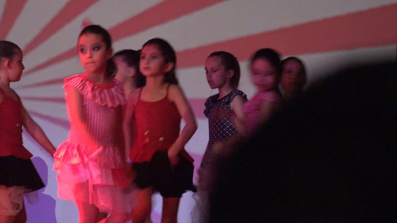 Several young girls face forward on a San Jose del Cabo BCS Mexico stage, with one girl in a pink ruffled costume near the front and others in red and polka-dot outfits behind her.