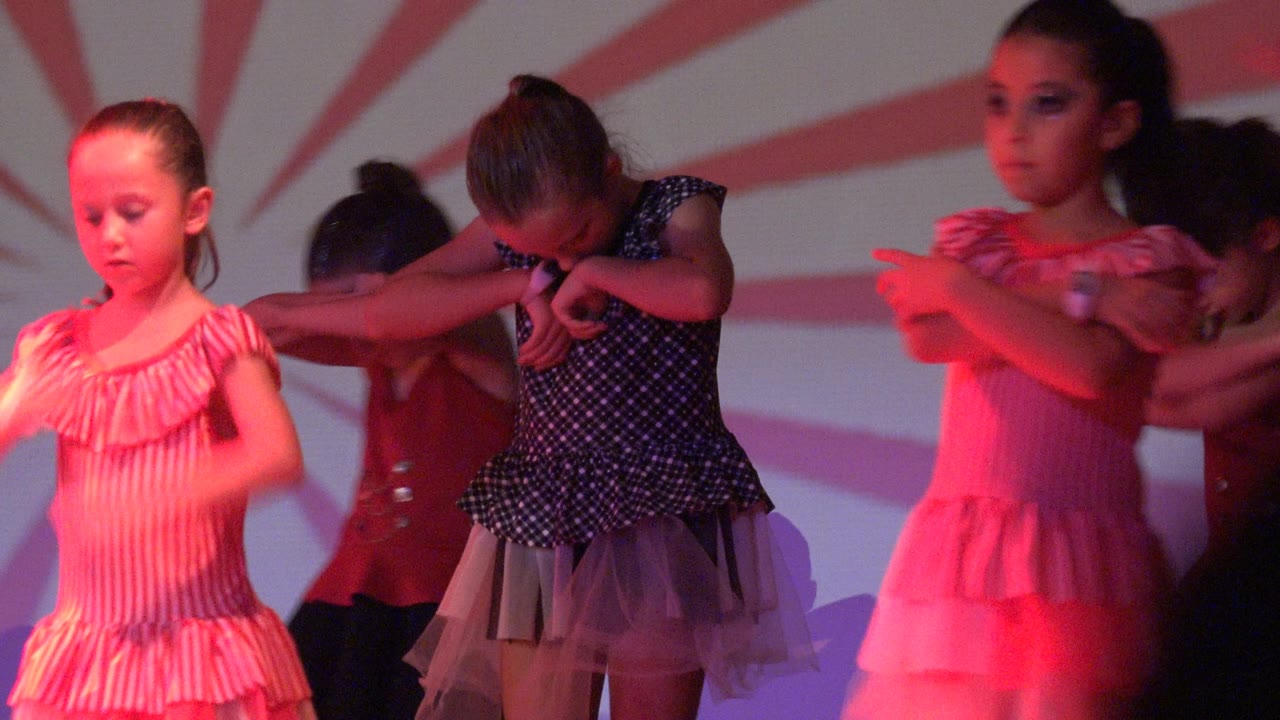 Young girls perform on a stage in Mexico, with a pink striped dress in front, a black polka-dot dress beside her, and red lighting across the dancers.