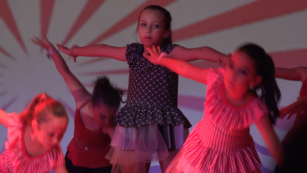 Several girls continue the dance on stage in Mexico, with one child in a pink striped dress facing forward and another in a black polka-dot dress turning sideways.