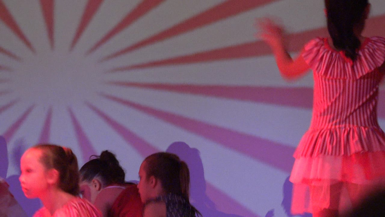 Young girls in colorful dresses are clustered low and upright on a red-lit stage in San Jose Del Cabo BCS Mexico, with a red and white sunburst backdrop behind them.
