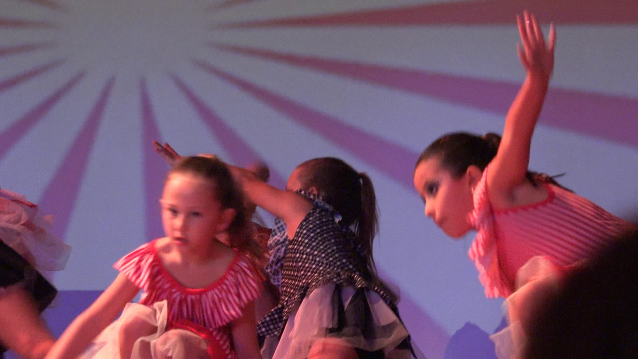 Girls perform arm movements in front of a sunburst backdrop