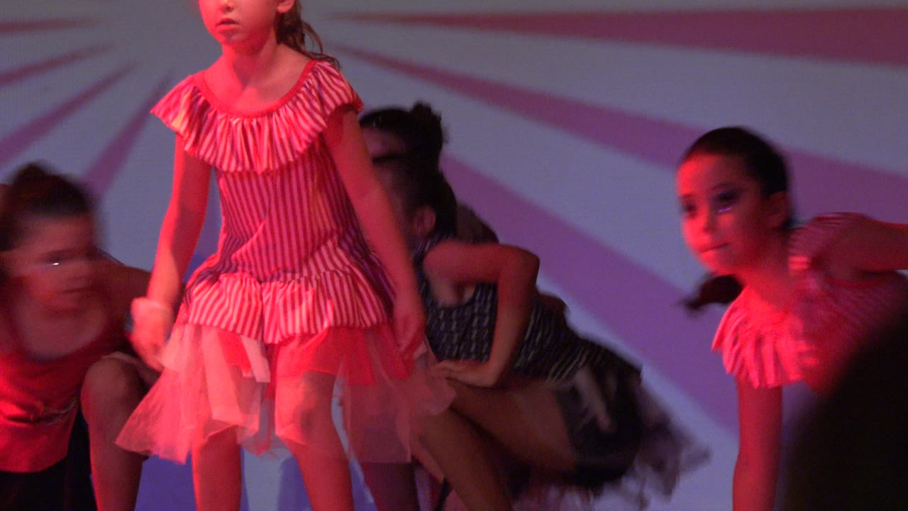Young dancers in striped and polka dot dresses occupy the stage in San Jose Del Cabo BCS Mexico, with one girl standing tall while others crouch behind her.