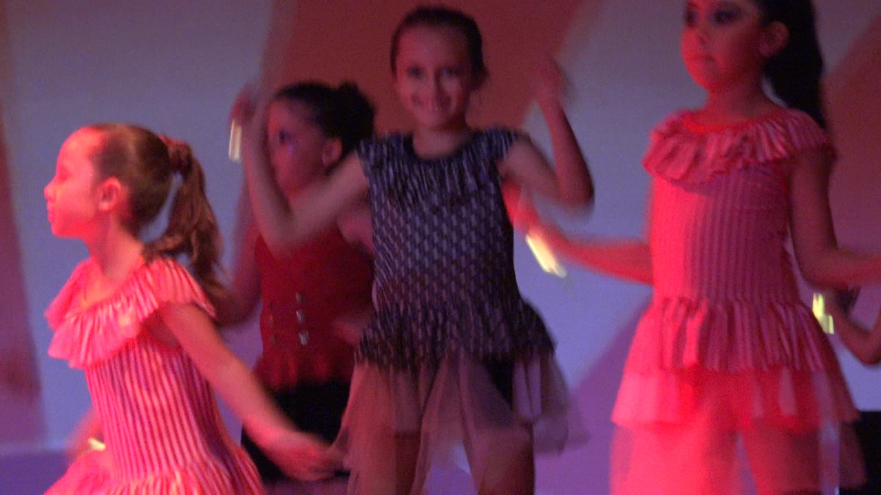 Young girls perform a dance routine on a stage in San Jose del Cabo BCS Mexico, with a child in a pink striped dress crossing the foreground and other dancers behind her.