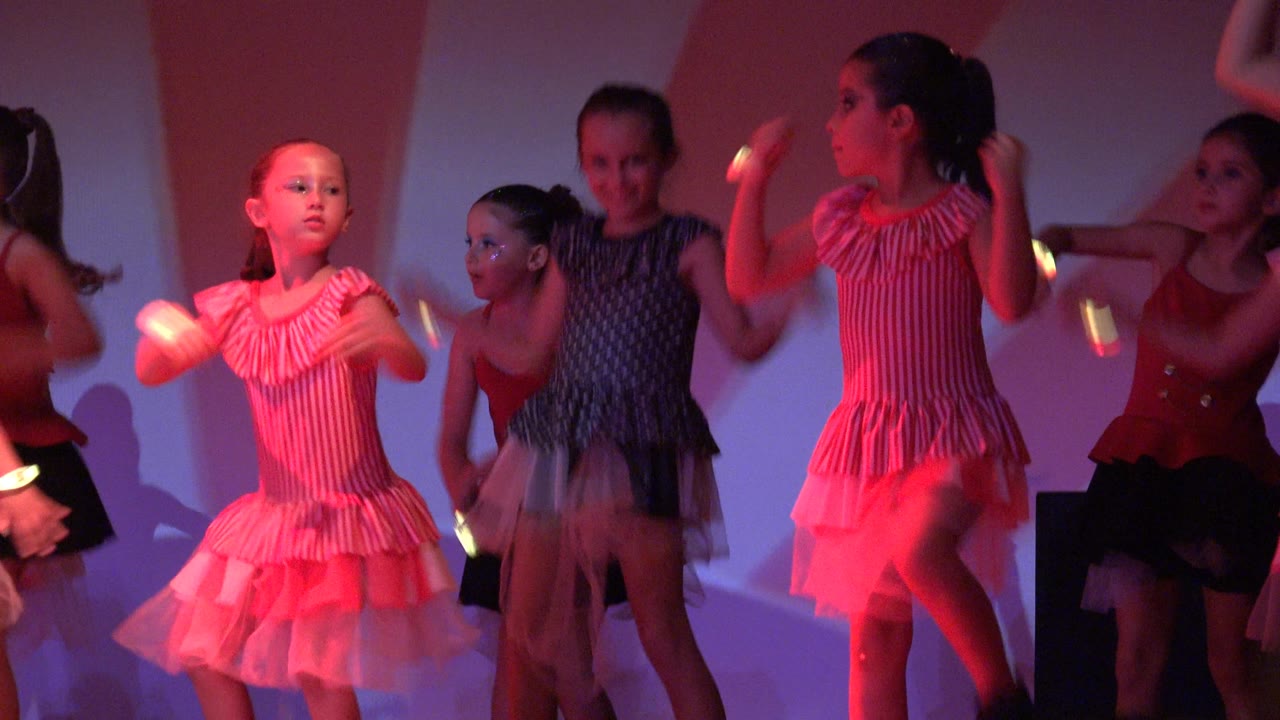 Young dancers in San Jose del Cabo BCS Mexico move through an arm gesture, with a girl in a pink striped dress in profile and other children partly visible behind her.