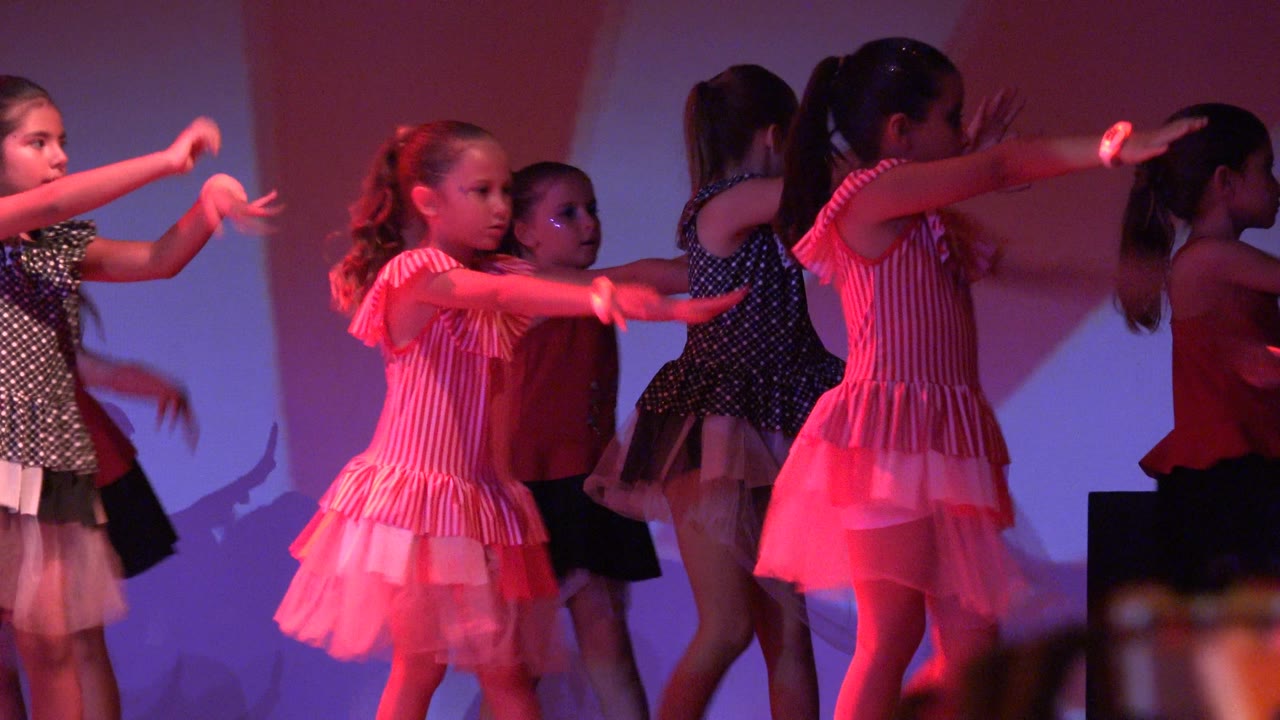 Young girls perform on a lit stage in San Jose Del Cabo BCS Mexico, with pink and red costumes, extended arms, and purple-red light across the backdrop.