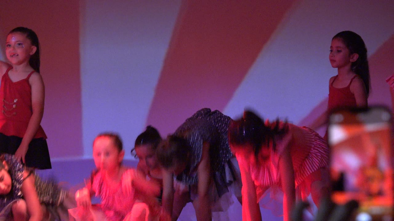 Young girls in red, striped pink, and dark polka-dot dance outfits perform on a stage in San Jose del Cabo BCS Mexico, with a blurred audience phone rising in the foreground.