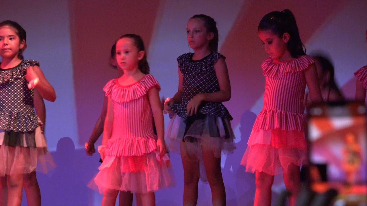 A girl in a pink striped tulle outfit and another in a dark polka-dot dress stand near center stage in San Jose del Cabo BCS Mexico as other children crouch around them.