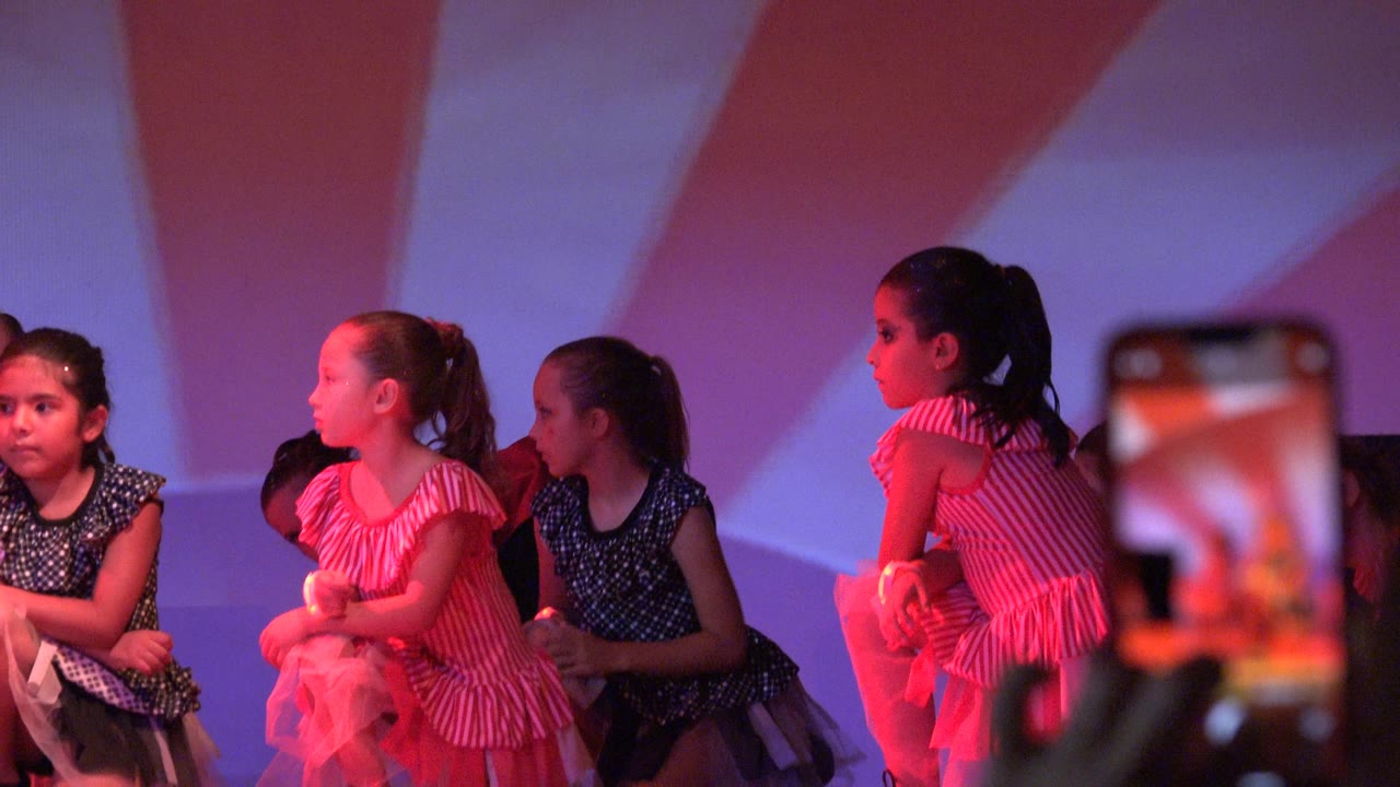 Young girls in pink ruffled and dark blue patterned dresses perform on a lit stage in San Jose Del Cabo BCS Mexico, with some dancers crouched and others facing sideways.