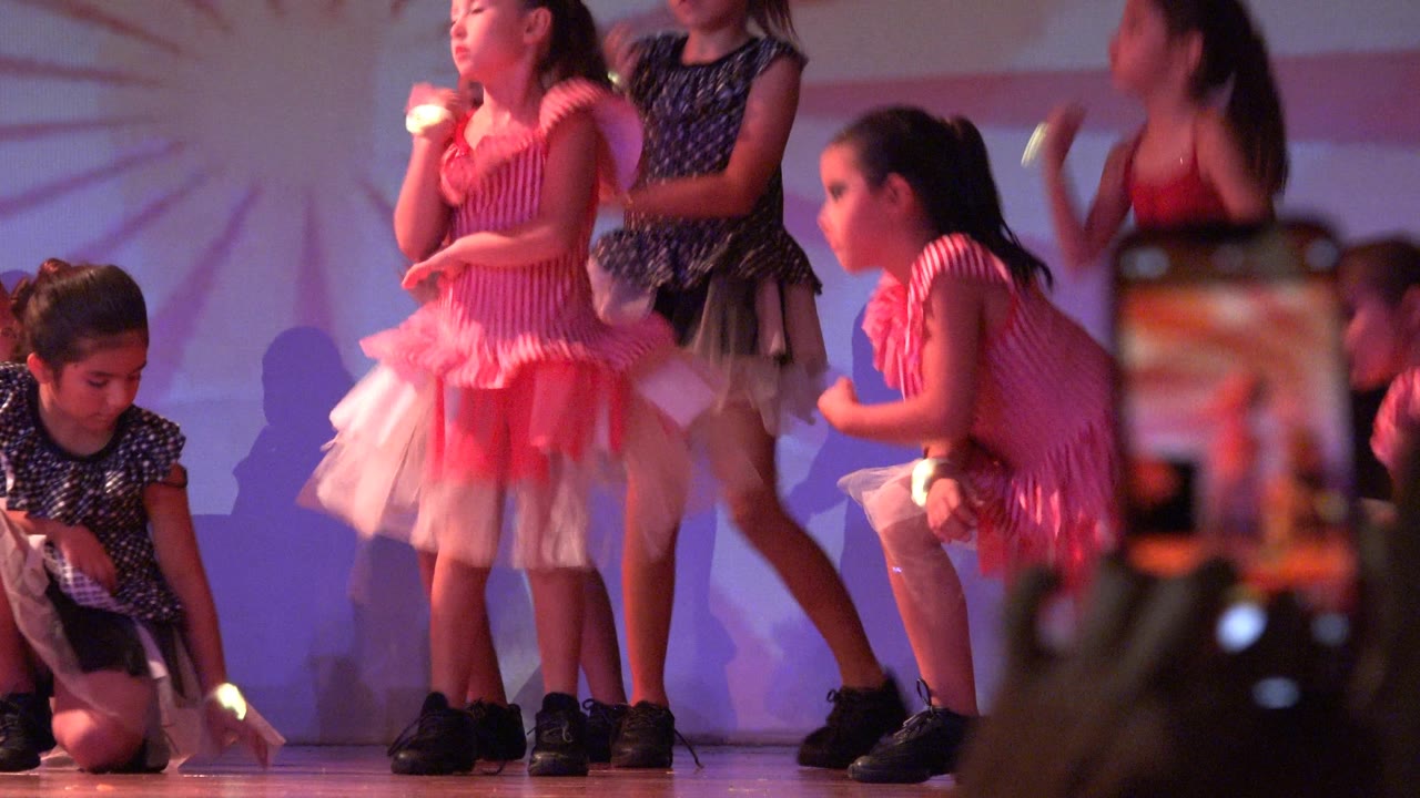 Children dance on stage while a smartphone records from the foreground.