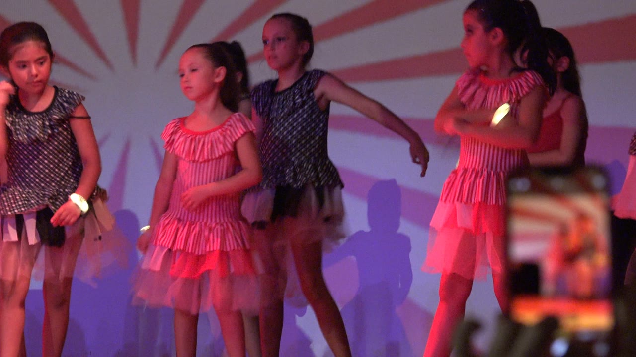 A line of young girls in pink, dark blue, and red stage outfits performs in San Jose Del Cabo BCS Mexico, with a blurred phone screen centered near the front of the frame.