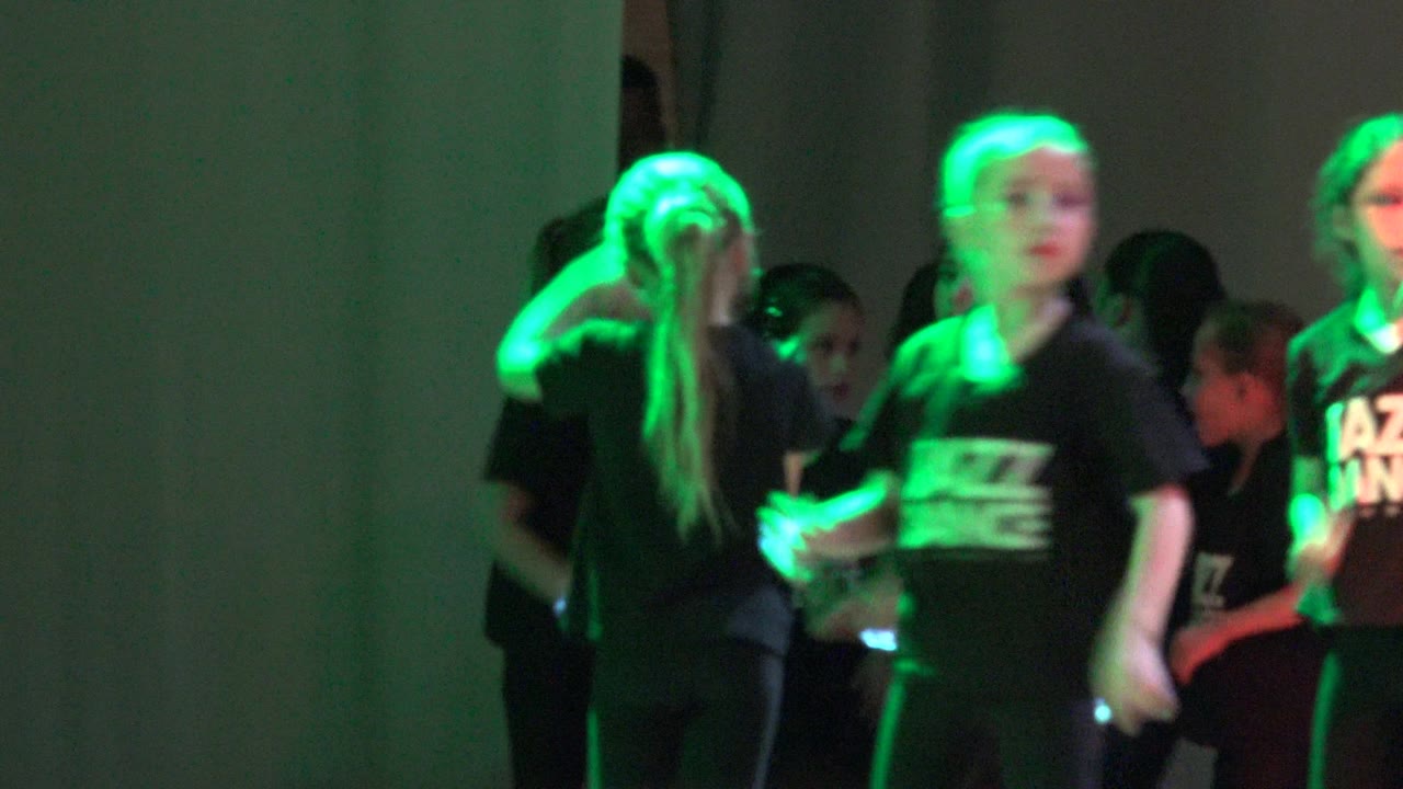 A child dancer stands near the front of the group in San Jose del Cabo BCS Mexico, with green light across the black Jazz Dance shirt and other children partly visible behind.