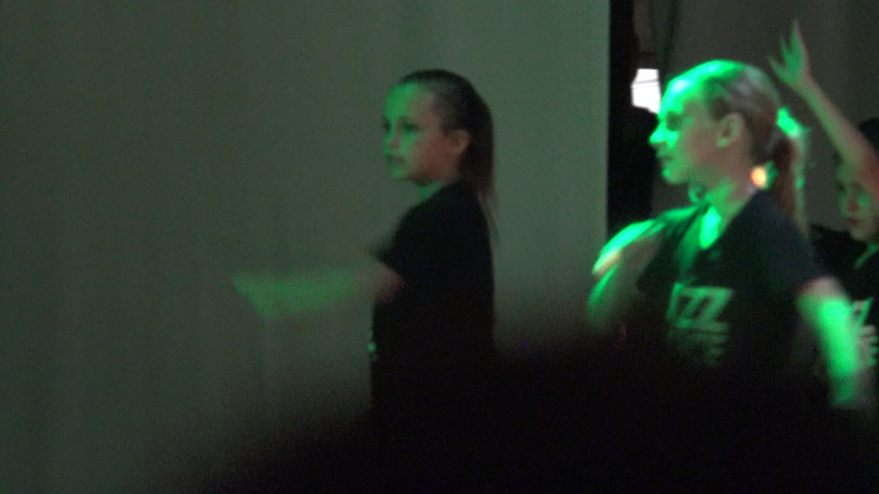 Children in black Jazz Dance shirts perform in San Jose del Cabo, Mexico as green light falls across their faces and a plain wall behind them.