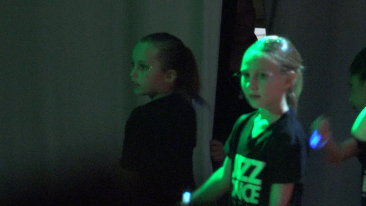 Children in San Jose Del Cabo, Mexico stand near a plain wall in a dim room as green glow-stick light falls across one girl's face and shirt.
