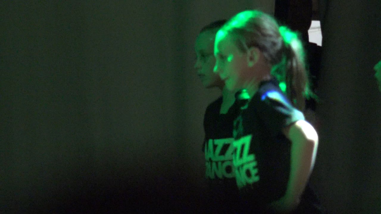 Children in black Jazz Dance shirts stand in profile in San Jose del Cabo, Mexico, with green stage light catching their hair and faces against a dark wall.