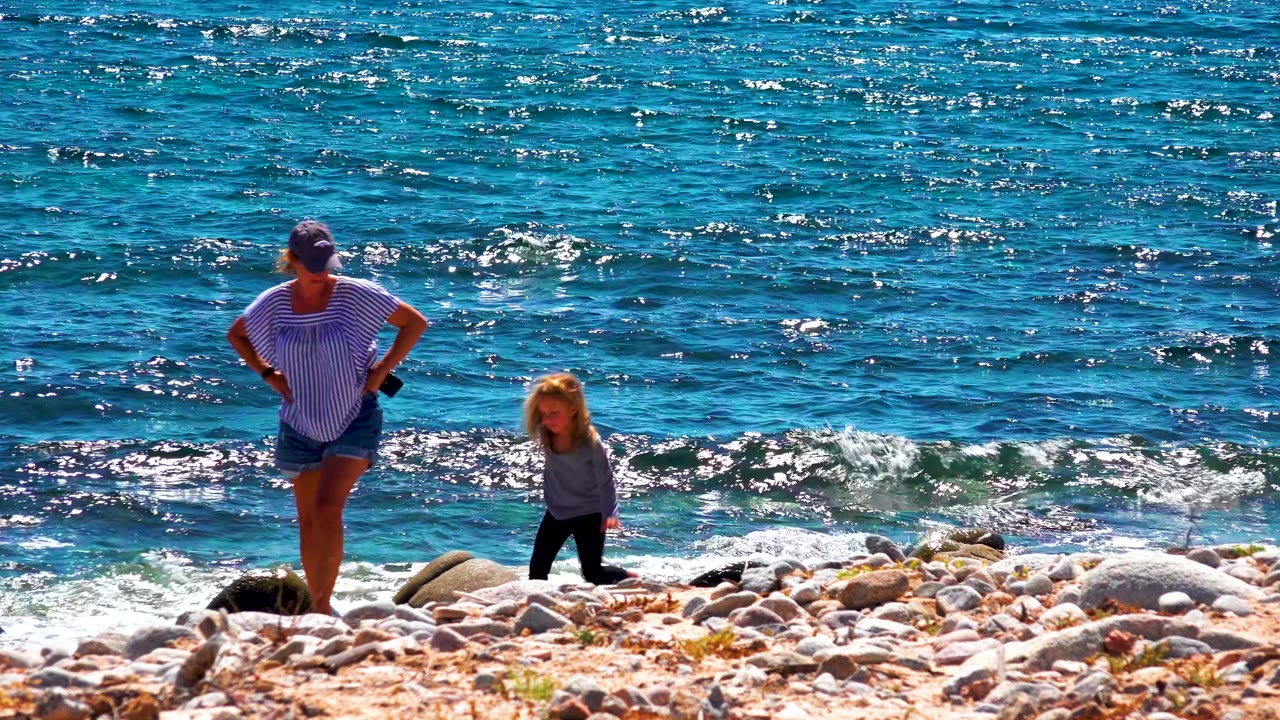 At El Sargento La Ventana Mexico, an adult in a striped shirt stands with hands on hips while a child walks across rocks near small waves.